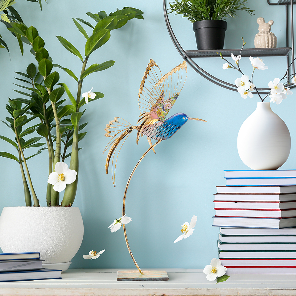 Elegant hummingbird figurine on a table with books and plants.